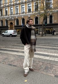 A man wearing a dark jacket, a checked scarf, and light trousers stands on a cobbled street in front of a yellow building with arched windows.