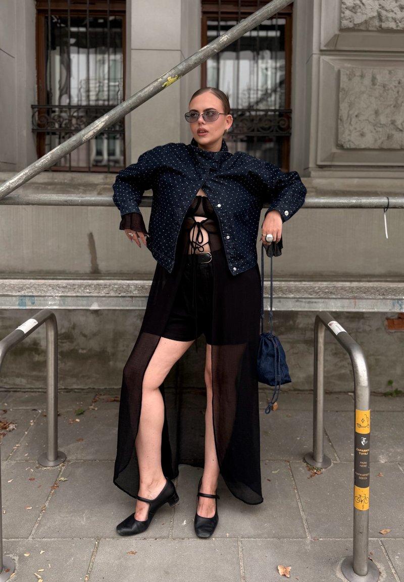 Personne portant une robe noire transparente avec un short en dessous, une veste sombre à pois, des lunettes de soleil et des chaussures noires, debout contre une rambarde métallique à l'extérieur.