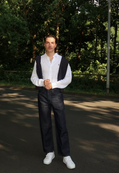 Camisa de lino blanca con mangas enrolladas, suéter azul marino colgado sobre los hombros, jeans azul oscuro y zapatillas blancas, de pie en una carretera rodeada de árboles.