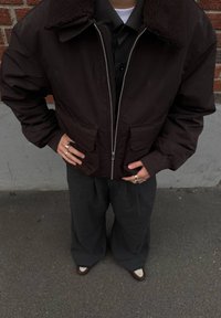 Giacca bomber marrone con colletto in pelliccia sintetica, chiusura con zip e due tasche frontali; abbinata a pantaloni grigi a gamba larga e scarpe marroni.