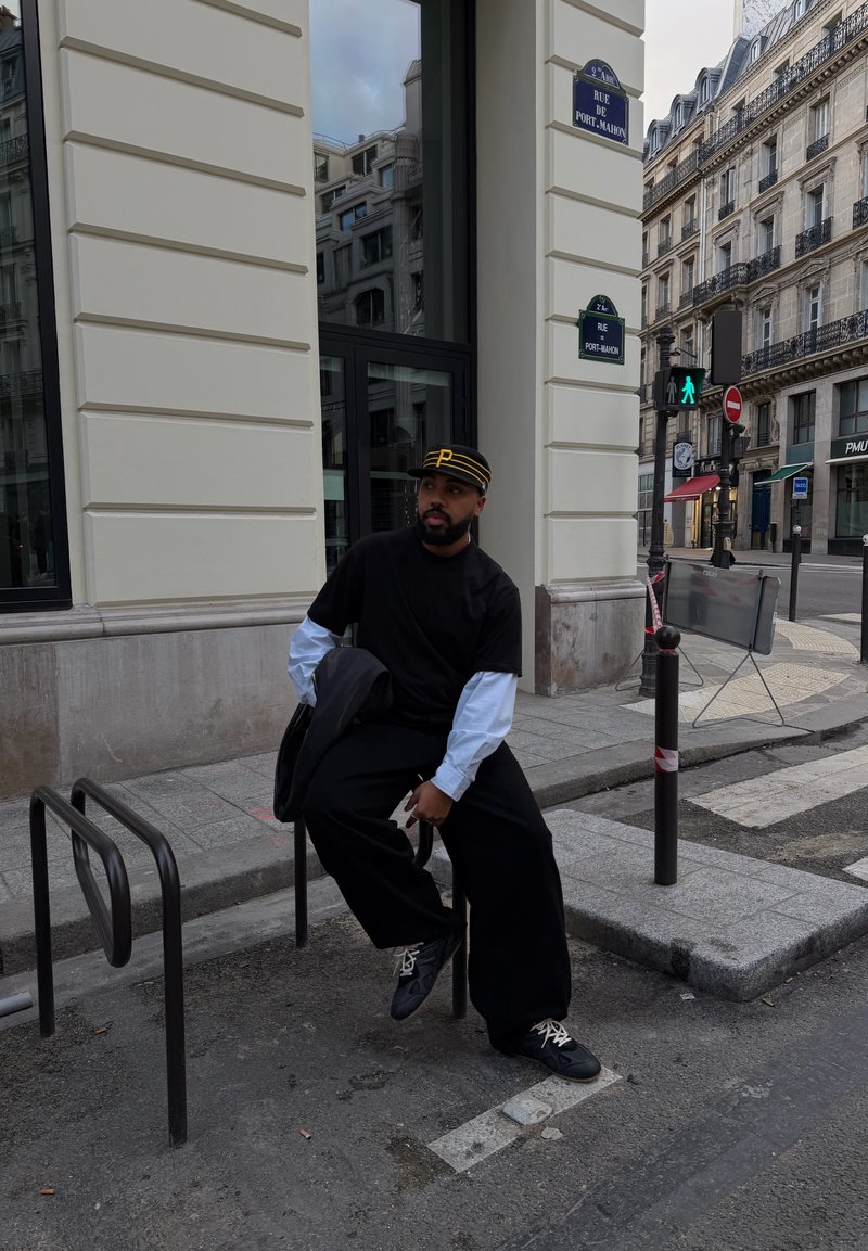 Homme portant des vêtements superposés noir et blanc et un chapeau noir, assis sur un support à vélos en métal à un coin de rue en ville.