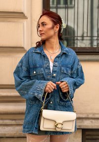 Nadrozmerná modrá džínsová bunda s prednými vreckami, biely top, krémová kabelka so zlatými detailmi a textúrované pančuchy; doplnky zahŕňajú zelenú sponu do vlasov a vrstvené náhrdelníky.