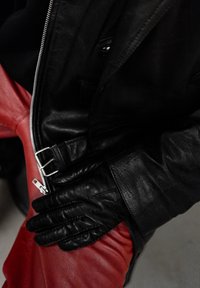 Des gants en cuir noir superposés sur une veste en cuir noir et un pantalon en cuir rouge vif, montrant des textures et des couleurs contrastées. Détail de la fermeture éclair visible.