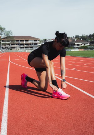 Mujer con ropa deportiva negra arrodillada en una pista de atletismo, atándose su zapatilla de correr rosa brillante, con edificios y zonas verdes al fondo.