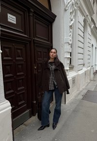 Caban en laine marron porté sur une chemise à carreaux, assorti à un jean bleu à jambes larges et à des bottines noires à bouts pointus. Un sac à main foncé repose sur le côté.