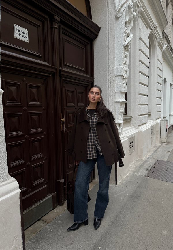 Caban en laine brune porté sur une chemise à carreaux, associé à un jean bleu à jambes larges et des bottines noires à bout pointu. Un sac à main sombre repose sur le côté.
