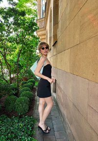 Μαύρο εφαρμοστό φόρεμα, ανοιχτές μαύρες γόβες, παστέλ μπλε τσάντα. Κοντά μαλλιά, υπέρμετρα γυαλιά ηλίου, πέτρινος τοίχος και πλούσια βλάστηση στο φόντο.