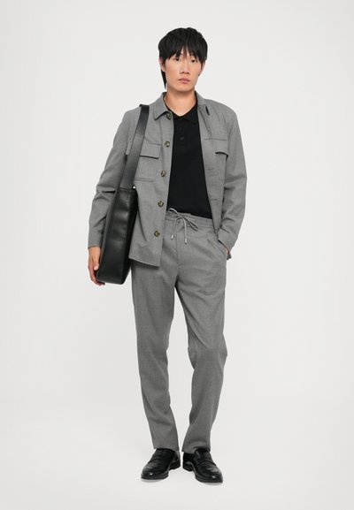 Traje gris a medida con chaqueta abotonada y pantalones con cordón. Camiseta polo negra debajo. Bolsa y zapatos de cuero negros.