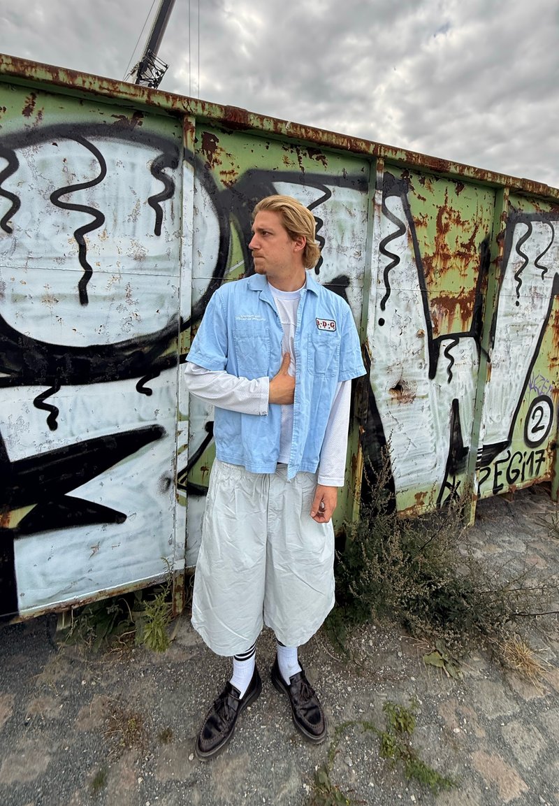 Lyseblå kortærmet skjorte og hvid mesh langærmet skjorte parret med knælange lyse shorts og mørke sko; graffiti baggrund.