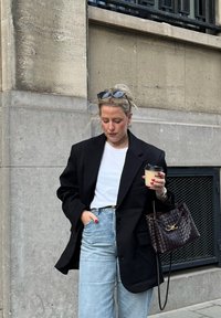 Femme portant un blazer noir surdimensionn é, un haut blanc, un jean bleu clair, tenant une tasse de café et un sac à main tressé sombre, debout près d'un mur en pierre en plein air.