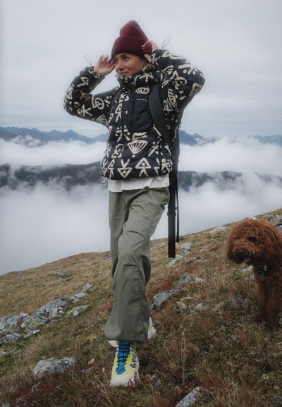 Chaqueta de plumas con un patrón en blanco y negro, pantalones verde olivo, zapatos de senderismo amarillos y azules, gorro de punto rojo, con un perro marrón cerca.