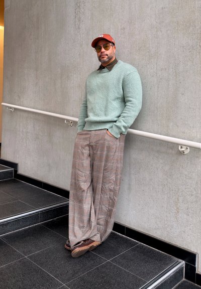 Hombre con gorra oxidada, gafas de sol amarillas, suéter verde menta, pantalones amplios de cuadros y zapatos marrones apoyado contra una pared de hormigón junto a unas escaleras.