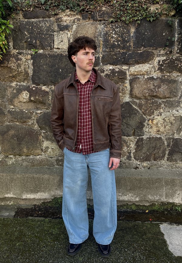 Rjava usnjena jakna čez rdečo in belo karirasto srajco, v kombinaciji s širokimi svetlo modrimi jeans-pantaloni in črnimi čevlji, pred kamenim zidom.