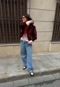 Femme portant des lunettes de soleil, une veste bordeaux, un pull rose, une écharpe rouge, un jean à jambes larges et des chaussures noir et blanc, debout sur le trottoir en ville.