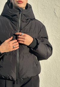 Doudoune noire avec un col montant, une fermeture éclair à l'avant et un petit détail de logo. Tissu texturé et poignets élastiques pour plus de chaleur.