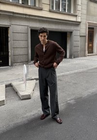 Bruine cardigan over een witte shirt, donkergrijze broek en bordeauxrode schoenen. Buiten in een stedelijke omgeving met gestructureerd asfalt en gebouwen.