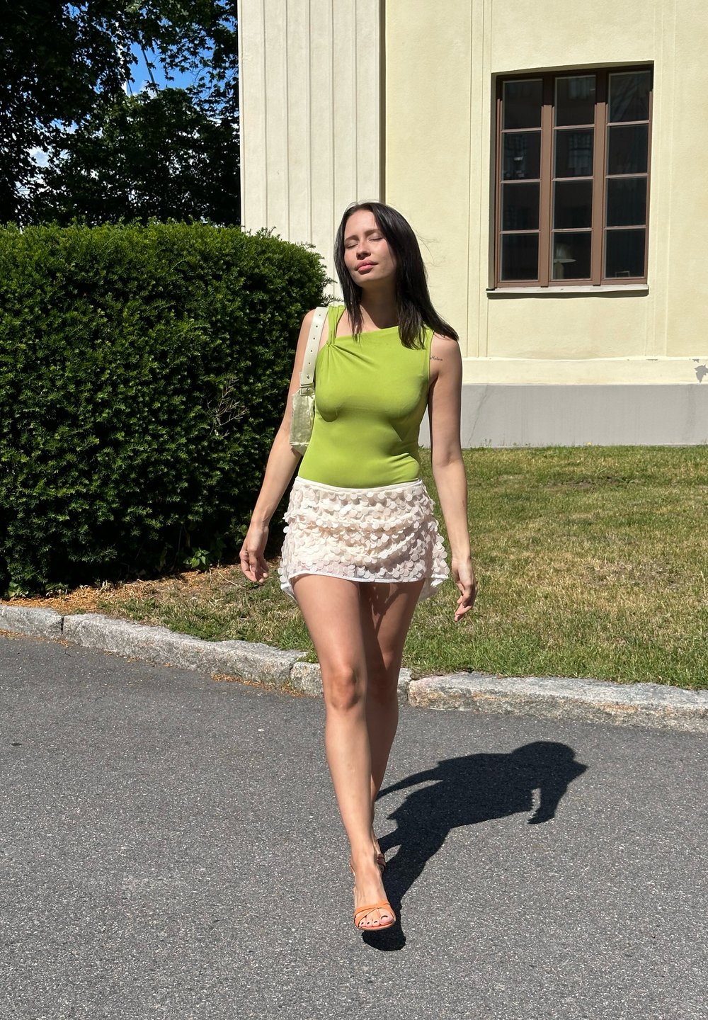 Zöld, ujj nélküli felső egy világos rózsaszín, fodros mini szoknyával és áttetsző táskával párosítva. Természetes környezet és napfény látható a háttérben.