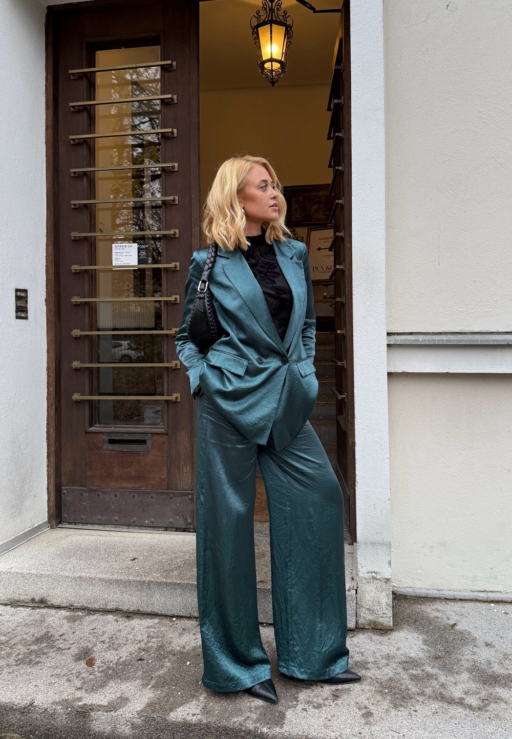 Tealfarget, strukturert dress med vide bukser og en skreddersydd jakke, kombinert med en svart topp og spisse svarte hæler, stående ved en tredør.