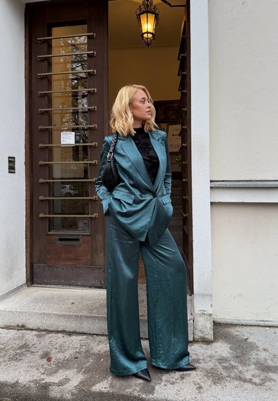 Traje verde azulado con textura, pantalones anchos y chaqueta entallada, combinado con una blusa negra y zapatos de tacón negros puntiagudos, de pie cerca de una puerta de madera.