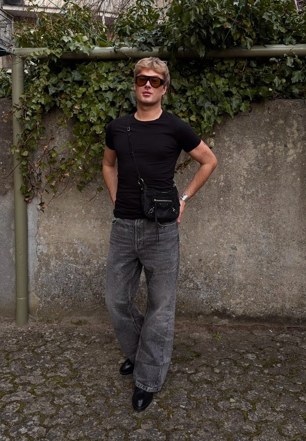 Tânăr purtând un tricou negru, blugi gri largi, cizme negre, ochelari de soare închiși la culoare și o geantă mică neagră purtată pe umăr, stând lipit de un zid de beton cu iederă.