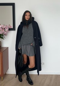 Palton lung bleumarin peste un pulover gri, fustă plisată gri și cizme negre până la genunchi. Accesorii include o geantă neagră. Fundal cu flori și oglindă.