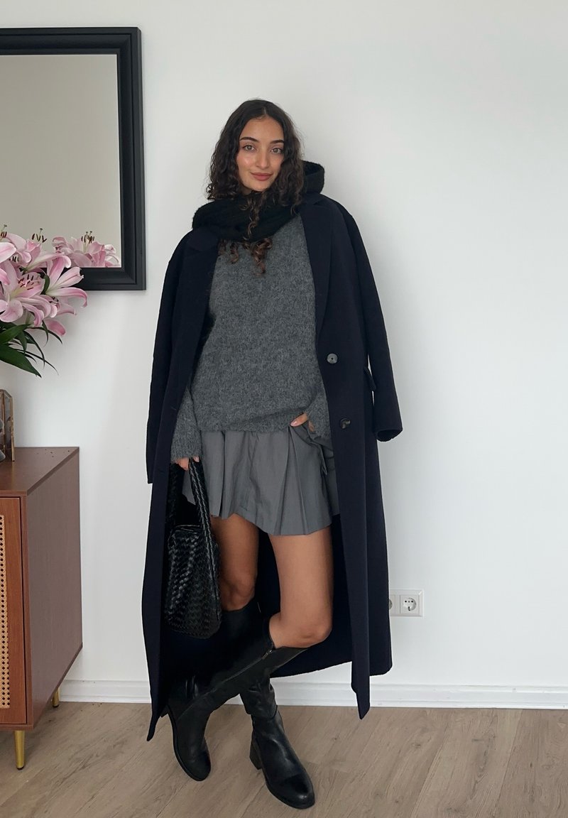 Lungo cappotto blu navy sopra un maglione grigio, gonna grigia plissettata e stivali neri al ginocchio. Accessori includono una borsa nera. Sfondo con fiori e specchio.