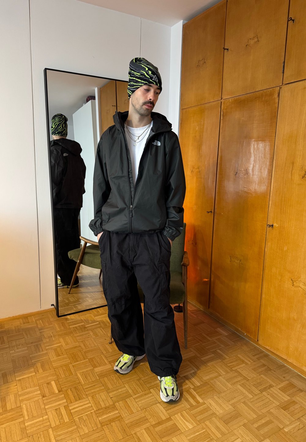 Homme portant une veste noire, un pantalon cargo noir, des baskets à accents néon et un bonnet noir et vert à motifs, debout à l'intérieur près d'un miroir.