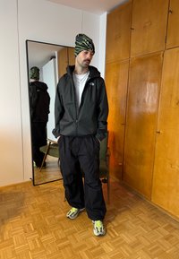 Homme portant une veste noire, un pantalon cargo noir, des baskets à accents néon et un bonnet noir et vert à motifs, debout à l'intérieur près d'un miroir.