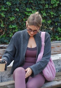 Grijze geribbelde cardigan, roze tanktop en bijpassende leggings; houdt een kopje koffie vast. Roze tas ligt naast haar. Groene bladeren op de achtergrond.