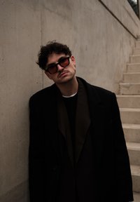 Homme aux cheveux bouclés et à la moustache, portant des lunettes de soleil foncées et un manteau, appuyé contre un mur en béton à côté d'un escalier en pierre.