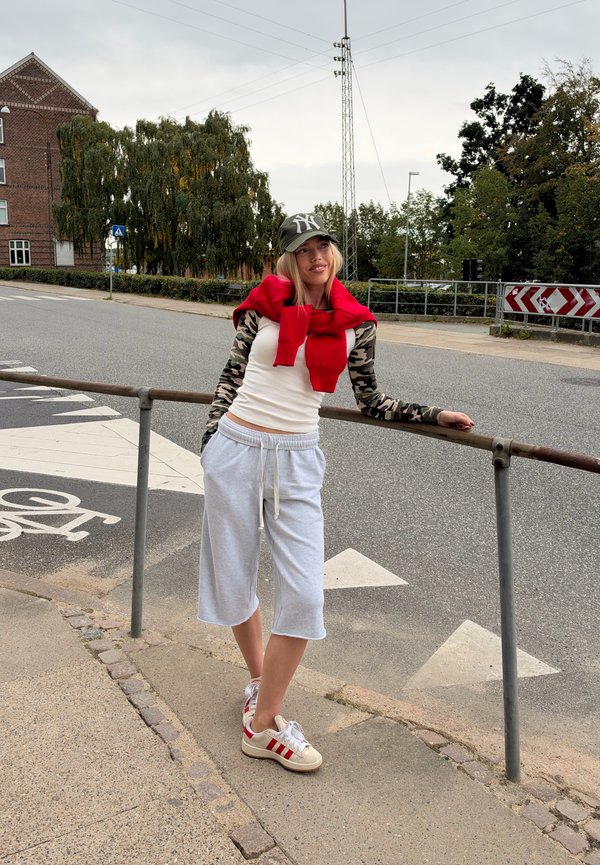 Una mujer con gorra de béisbol, jersey rojo y pantalones cortos grises y holgados se apoya en una barandilla junto a un paso de peatones.