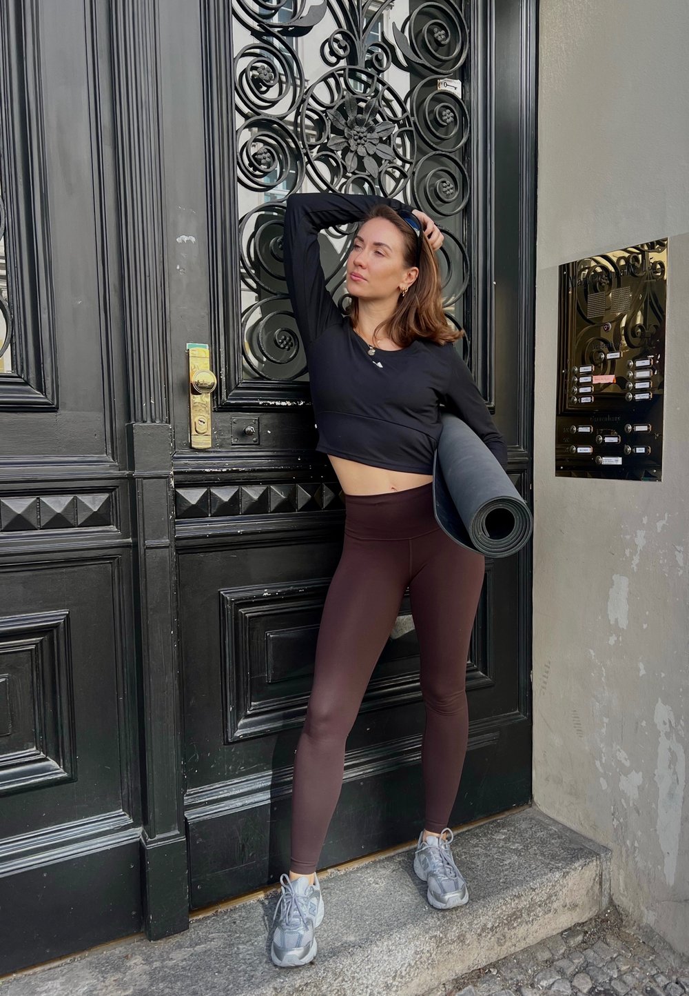 Black long-sleeve crop top, burgundy leggings, and grey sneakers. Rolled yoga mat held in one arm. Intricate black door in the background.