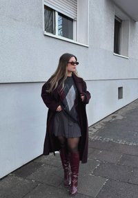 Femme portant des lunettes de soleil, un long manteau sombre, un pull à losanges, une jupe plissée, des collants noirs et des bottes bordeaux montant jusqu'aux genoux, marchant sur un trottoir.