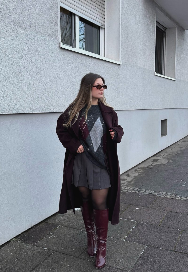 Femme portant des lunettes de soleil, un long manteau sombre, un pull à losanges, une jupe plissée, des collants noirs et des bottes bordeaux montant jusqu'aux genoux, marchant sur un trottoir.