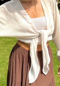 Kobieta w białej bluzce zawiązanej z przodu, założonej na biały, szydełkowany krótki top i brązową plisowaną spódnicę, stojąca na trawie na zewnątrz.