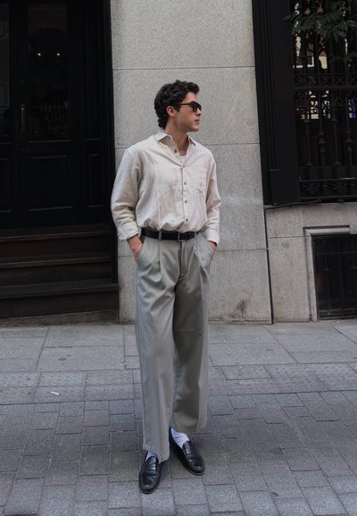 Hombre con pelo oscuro que lleva gafas de sol, una camisa clara de rayas, pantalones anchos grises y zapatos negros, de pie con las manos en los bolsillos en una acera de la ciudad.
