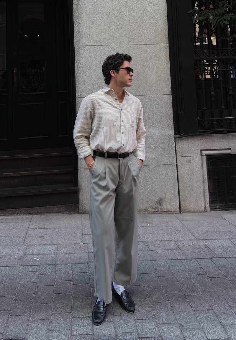 Homme aux cheveux foncés portant des lunettes de soleil, une chemise claire à rayures, un pantalon large gris et des chaussures noires, se tient les mains dans les poches sur un trottoir en ville.