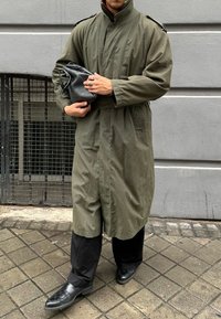 Ελιά πράσινο, παλτό μέχρι το γόνατο με ψηλό γιακά και επωμίδες στους ώμους, συνδυασμένο με μαύ�ρα παντελόνια και γυαλιστερές μαύρες γόβες. Μαύρη τσάντα στο χέρι.