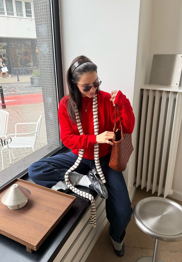 Mujer con suéter rojo y gafas de sol sentada junto a una ventana mirando dentro de un bolso marrón tejido con las piernas cruzadas y zapatos casuales.