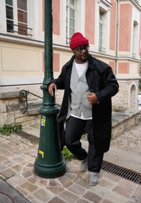 Hombre con gorro rojo, gafas, cárdigan gris, abrigo negro y vaqueros apoyado en un farol pintado de verde oscuro en una acera de adoquines.