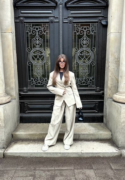 Mujer con cabello largo, vistiendo un traje beige y zapatos blancos, sosteniendo un bolso negro, de pie sobre escalones de piedra frente a unas rejas de metal negro ornamentadas.