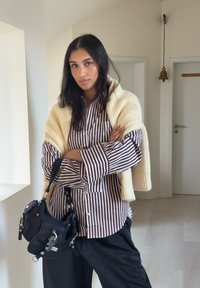 Geantă neagră cu accente metalice, purtând o cămașă în dungi cu dungi bej și un șal tricotat crem. Pantaloni largi negri uni.