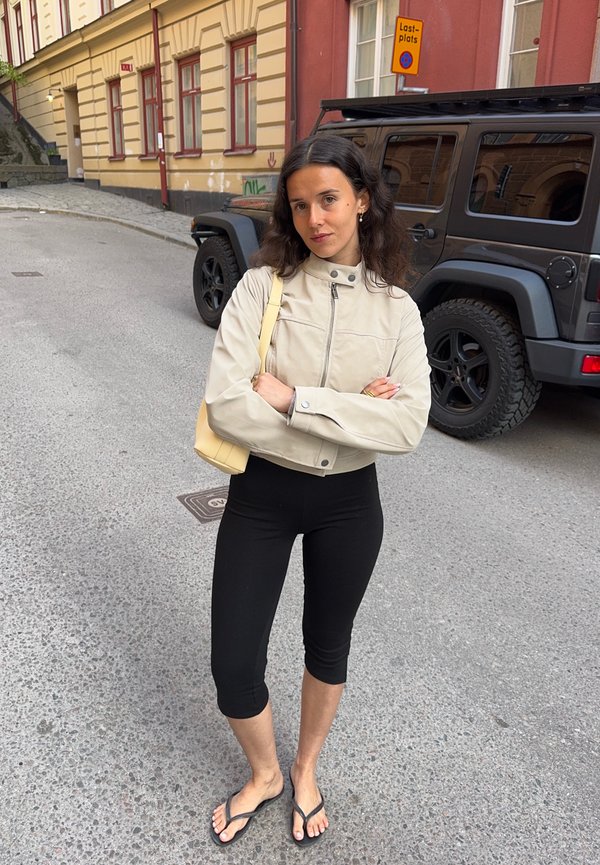 Casaco cropped bege com gola alta e fecho à frente, leggings capri justas pretas e chinelos pretos. Saco de ombro amarelo claro, em ambiente de rua.