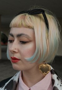 Kort bob-frisyre med pastellrosa og lys blå nyanser. Kjennetegnes av hjerteformede gulløredobber, fremtredende nesepiercinger og dristige røde lepper.