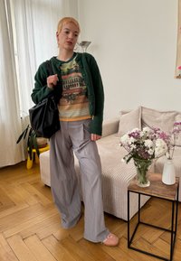 Cardigã verde de lã, suéter com padrão gráfico, calças largas cinzas, bolsa preta oversized, chinelos rosas. Ambiente interior simples com plantas.