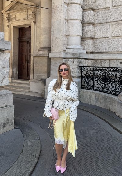 Mujer vistiendo blusa de lunares, falda amarilla, tacones rosas y gafas de sol, de pie sobre un pavimento de piedra cerca de la entrada ornamentada de un edificio de piedra.