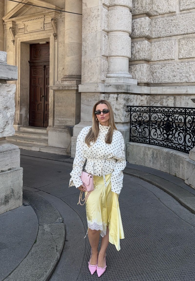 Femme portant un chemisier à pois, une jupe jaune, des talons roses et des lunettes de soleil, debout sur un pavé en pierre près d'une entrée richement ornée en pierre d'un bâtiment.