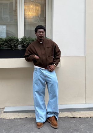 Chaqueta bomber de forro marrón con bolsillos frontales, jeans sueltos de color azul claro y zapatos de ante marrones. De pie contra una pared blanca con vegetación.