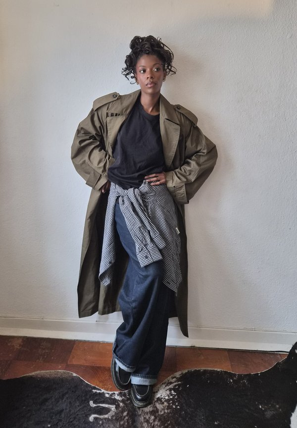 Une femme pose devant un mur uni, vêtue d’un trench olive, d’une chemise noire, d’un jean à jambes larges et de chaussures noires. Une chemise à carreaux est nouée autour de sa taille.