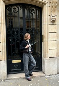 Zwarte stoffen jas met gouden knopen, witte coltrui, grijze wijde broek, en bordeauxrode schoenen, staand bij een opvallende zwarte ijzeren deur.